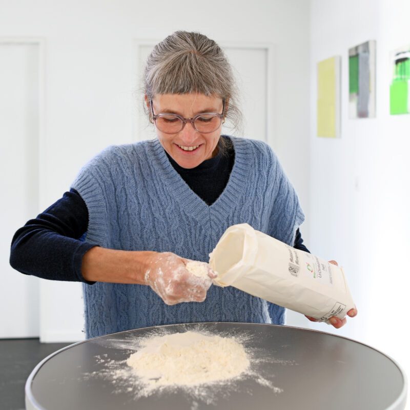 Silvia Bolliger hat eine Mehltüte in der Hand und streut mit der anderen Hand Mehl auf den Tisch vor sich.