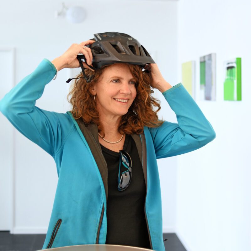 Doris Jost setht sich lächelnd einen Velohelm auf. Sie trägt eine türkisfarbene Jacke und hat eine Sonnenbrille beim T-Shirt Kragen eingehängt.