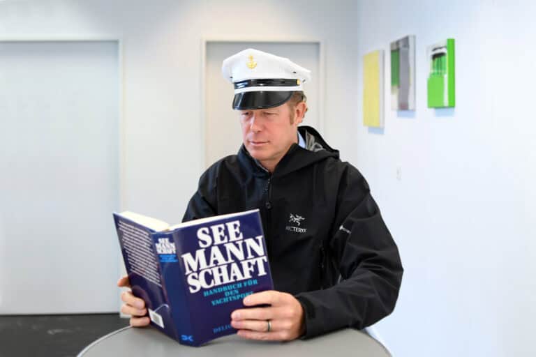 Michael Kiehl trägt eine weisse Kapitänsmütze und eine blaue Jacke im Marine Stil. Er liest n einem Handbuch für den Yachtsport.