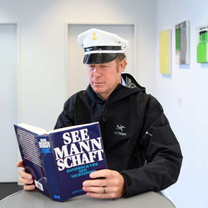 Michael Kiehl trägt eine weisse Kapitänsmütze und eine blaue Jacke im Marine Stil. Er liest n einem Handbuch für den Yachtsport.