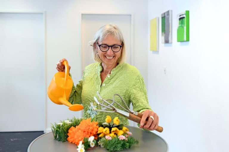 Barbara Krieger stet an einem Tisch mit einem farbigen Blumenarrangement aus künstlichen Blumen, in der rechten Hand eine gelbe Spritzkanne und in der linken hand zwei Gartengeräte