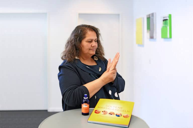 Natalie Mollah steht an einem Tisch und massiert sich die Hand, vor sich eine Flasche mit Massageöl und ein Buch mit den Goldenen Massageregeln.