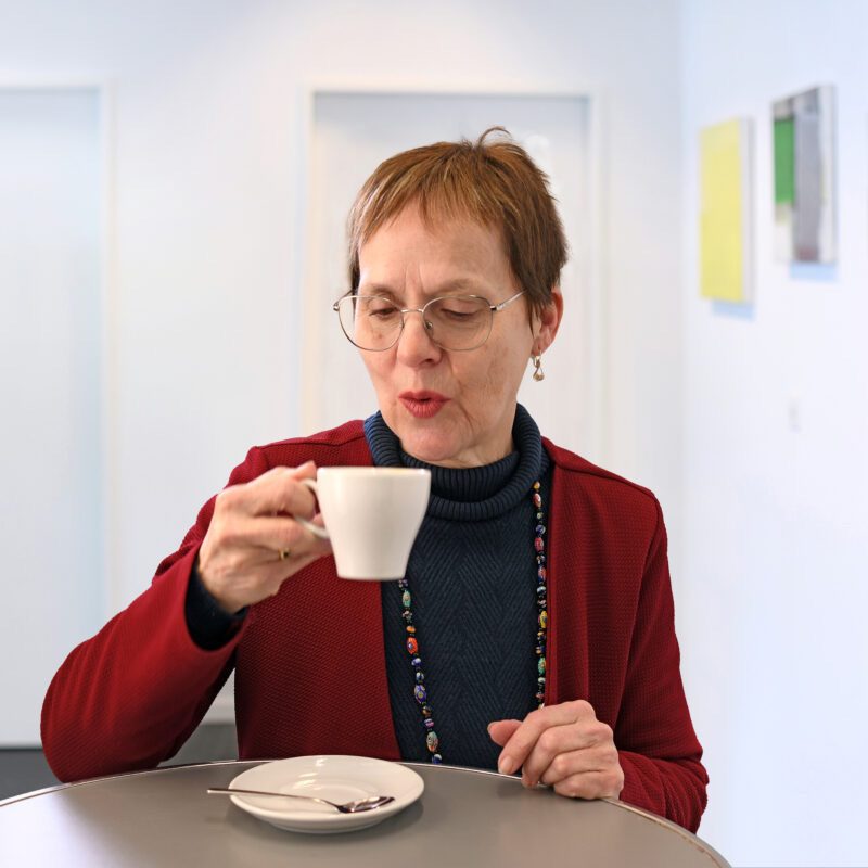 Beatrice Schneider steht an einem Tisch mit einer Tasse in der Hand. Der Kaffee darin ist offenbar noch heiss, sie bläst in die Tasse um den Kaffee zu kühlen.